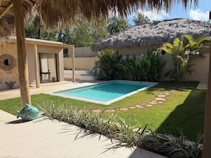 Ein Pool mit einer strohgedeckten Struktur, umgeben von einem gepflegten Rasen und tropischen Pflanzen.