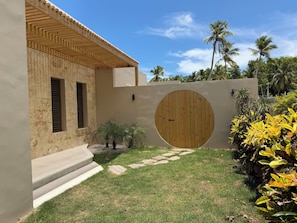 A modern house with a wooden door, a stone wall, and a small garden with palm trees. - Brand-New Luxury Villa · Private Pool · 5-Min Walk to Playa Cosón (Las Terrenas)