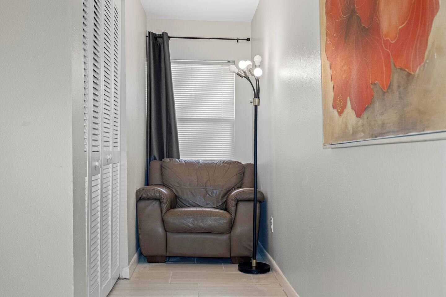 Ein schmaler Flur mit einem Ledersofa, einer Stehlampe mit mehreren Leuchtmitteln, einem Fenster mit Jalousien und einem teilweisen Blick auf ein Bild an der Wand.