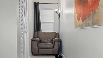 Ein schmaler Flur mit einem Ledersofa, einer Stehlampe mit mehreren Leuchtmitteln, einem Fenster mit Jalousien und einem teilweisen Blick auf ein Bild an der Wand.