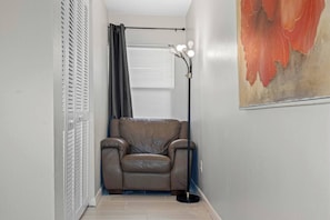 Ein schmaler Flur mit einem Ledersofa, einer Stehlampe mit mehreren Leuchtmitteln, einem Fenster mit Jalousien und einem teilweisen Blick auf ein Bild an der Wand.