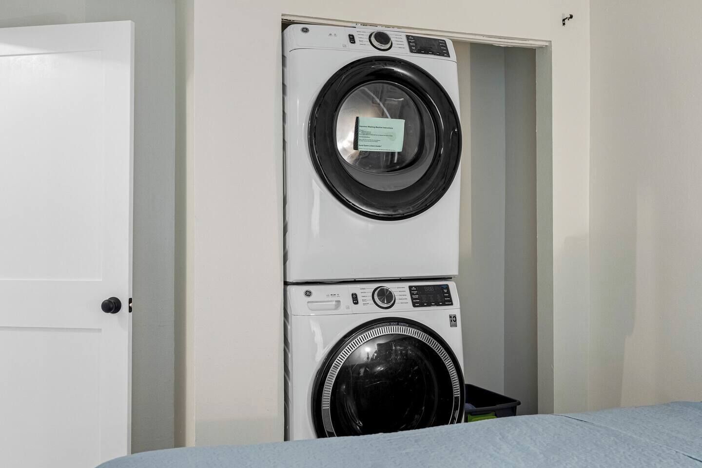 Eine übereinander gestellte Waschmaschine und ein Trockner in einem Raum mit einer weißen Tür und einem Bett mit blauer Bettdecke.