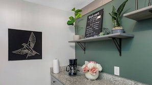 A kitchen with a coffee maker, a vase of flowers, a chalkboard with a welcome message, and a wall-mounted bird illustration. - Magnolia House; Grille/Firepit/Patio View! (Tampa)