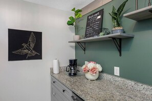 Eine Küche mit Kaffeemaschine, Blumen in einer Vase, einer Tafel mit einer Willkommensbotschaft und einer an der Wand befestigten Vogelillustration.