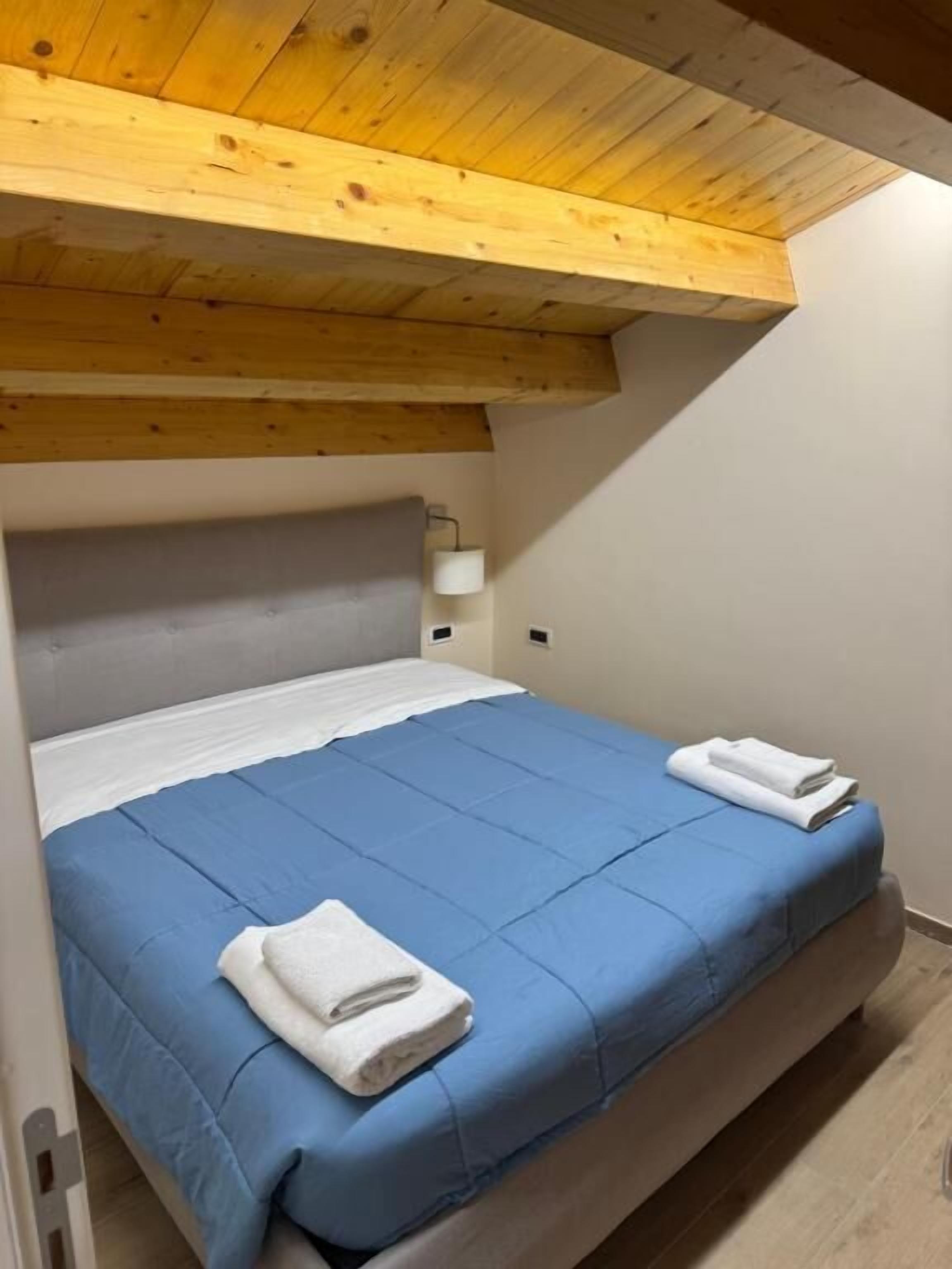 A single bed with a blue mattress and white linens, a wooden ceiling, and a white wall.