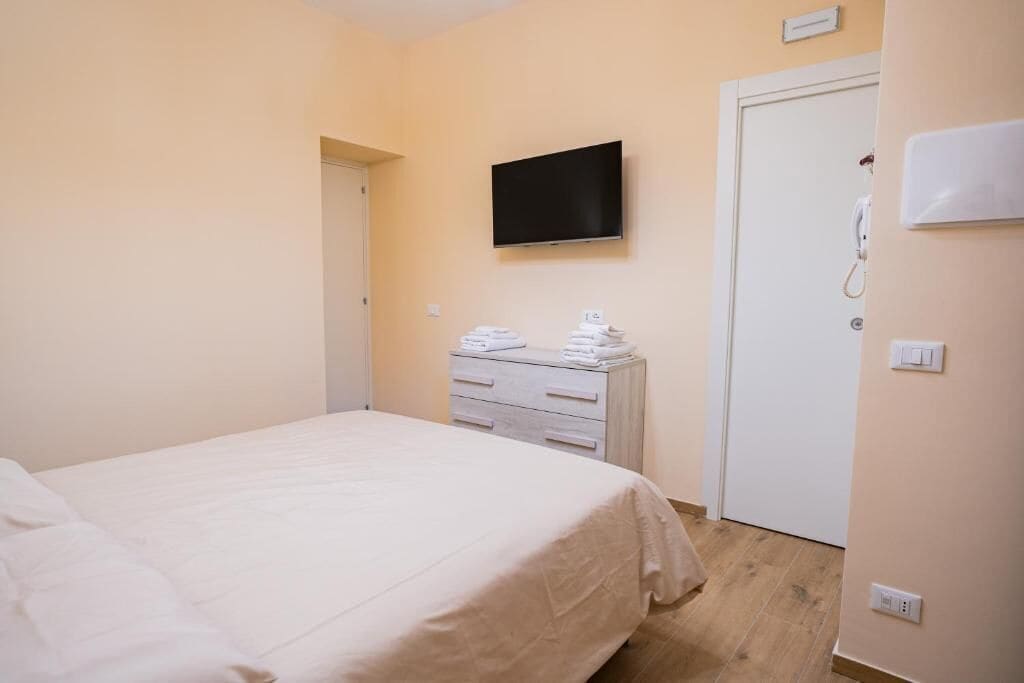 Une chambre d’hôtel avec un lit, un téléviseur fixé au mur, une table de chevet avec des serviettes et une porte avec un téléphone.
