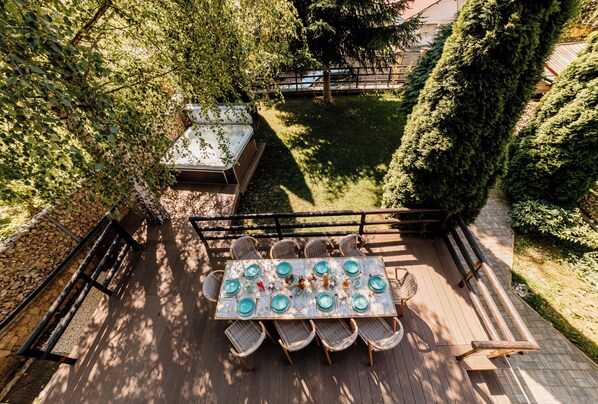 An outdoor dining area with a table set for six, surrounded by chairs, on a wooden deck with a view of a garden and a hot tub.