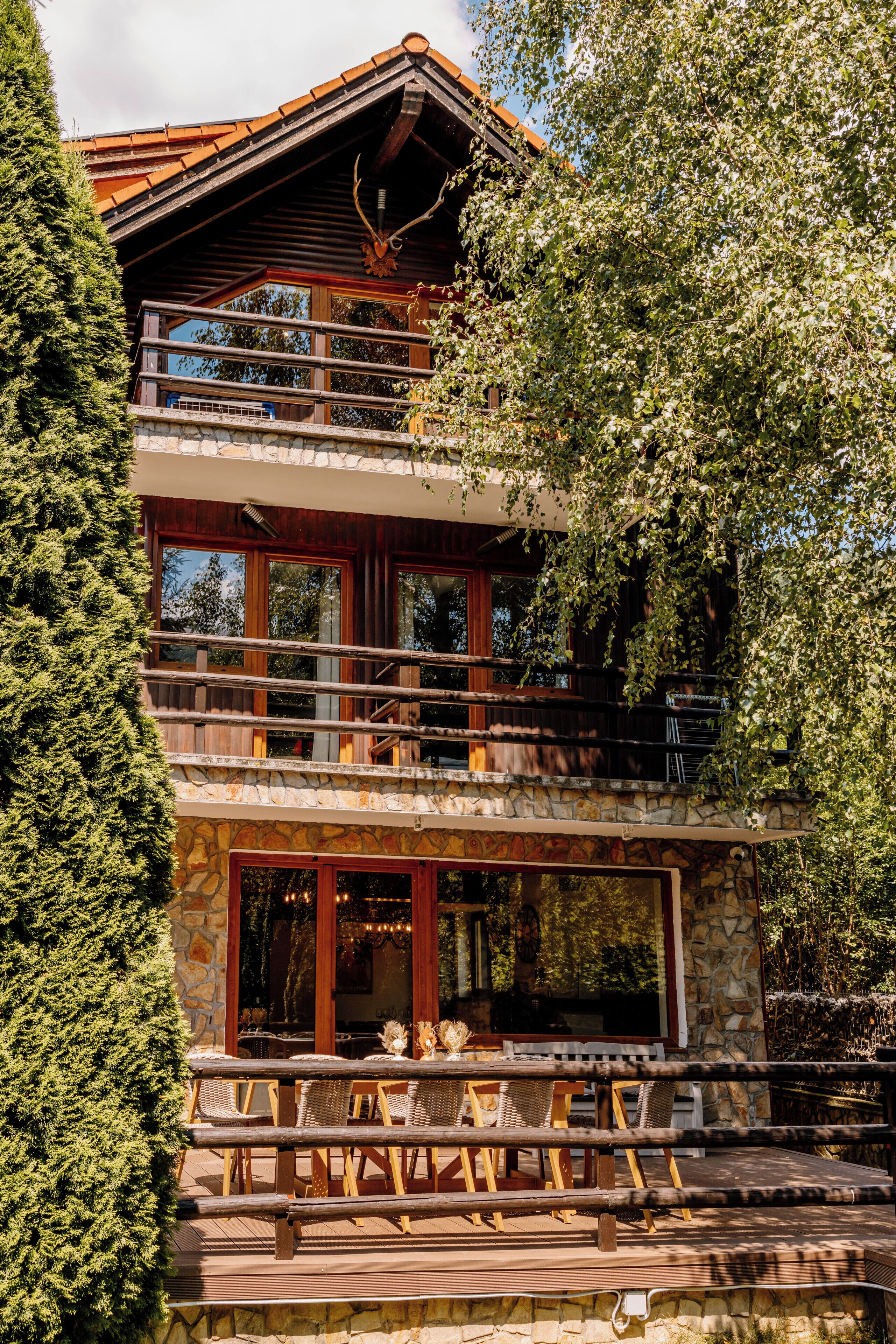 A multi-level wooden cabin with a stone facade, large windows, and a balcony with wicker furniture.