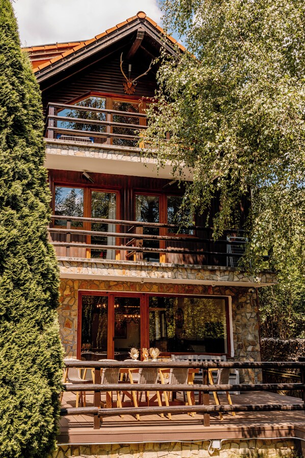 A multi-level wooden cabin with a stone facade, large windows, and a balcony with wicker furniture.
