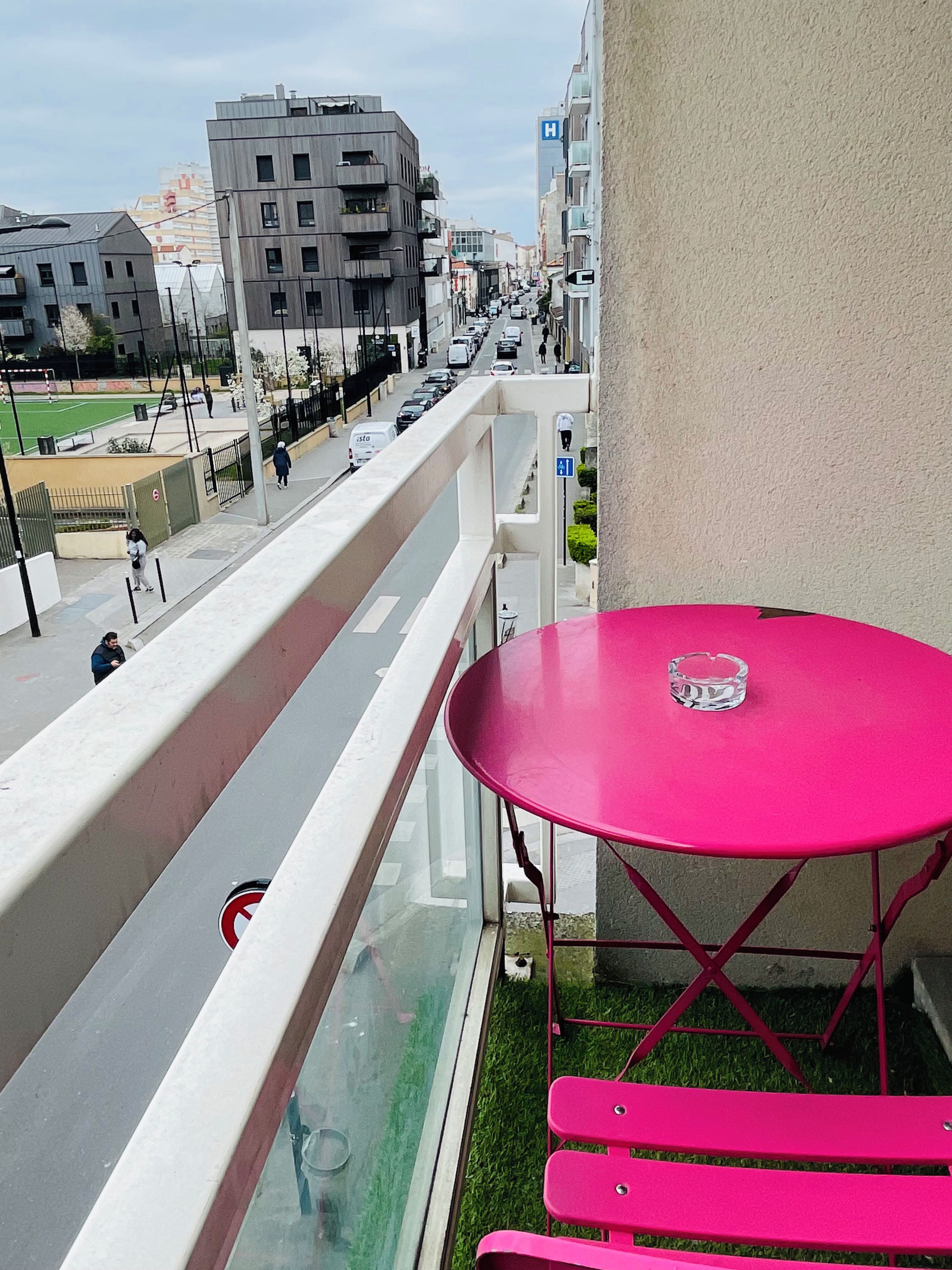 Un balcone con un tavolo e panche rossi, che si affaccia su una strada cittadina con edifici e auto parcheggiate.