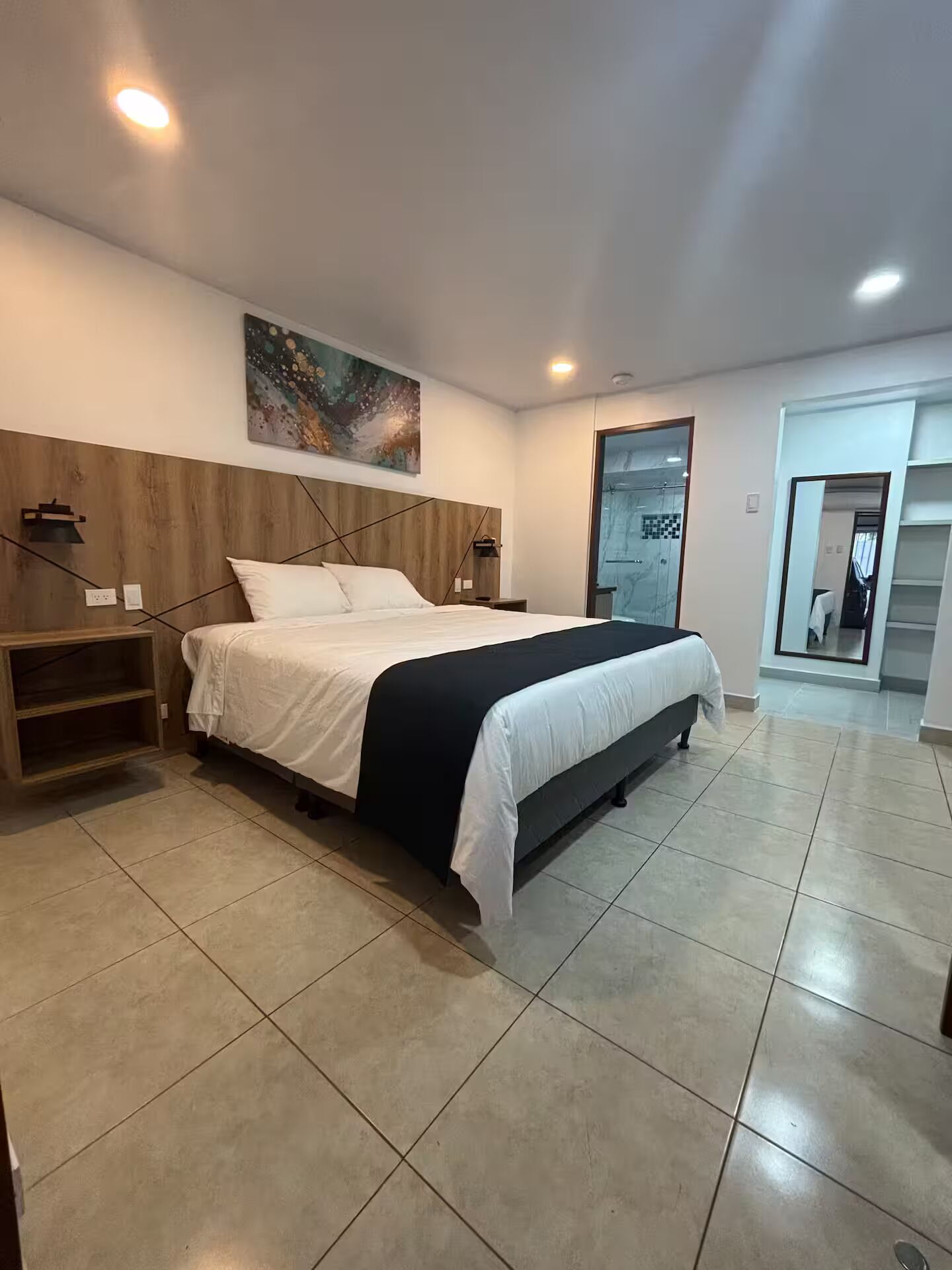 A modern bedroom with a large bed, a wooden headboard, a painting on the wall, and a door leading to another room.