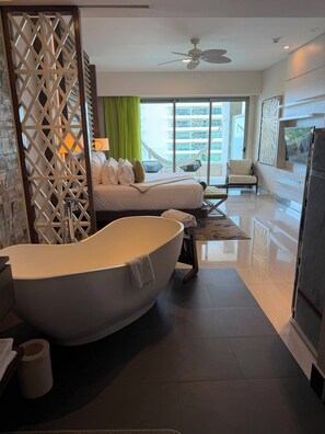 Ein modernes Hotelzimmer mit einer großzügigen Badewanne, einem Bett mit Kissen, einer Sitzecke mit einem Sessel und Blick ins Freie.