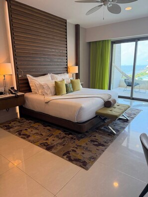 Een moderne hotelkamer met een groot bed, een nachtkastje met een lamp, uitzicht op zee door een groot raam, en een plafondventilator.