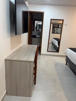 A modern hotel room with a wooden dining table, a bed, a mirror, and a view into the kitchen area.