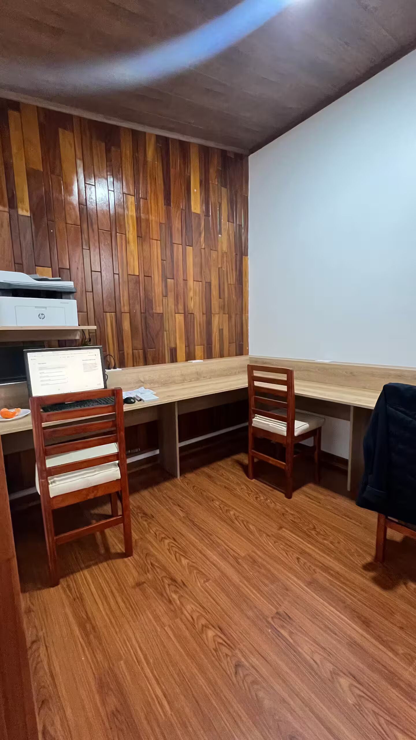 Um quarto com piso de madeira, uma escrivaninha com impressora, duas cadeiras e uma lousa.