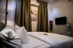 A hotel room with a bed, curtains, a wall-mounted TV, a vase with flowers, and a standing fan. - 10 minutes from Airport | One-Bed Luxury Stay in Lagos (Ikeja)