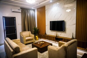 A modern living room with a flat-screen TV mounted on the wall, a sofa set, a coffee table, and a decorative plant. - 10 minutes from Airport | One-Bed Luxury Stay in Lagos (Ikeja)