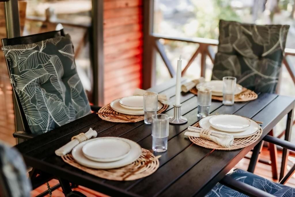 A dining table set with plates, glasses, and a candle, surrounded by chairs with patterned cushions.