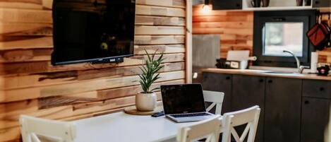 Una habitación con pared de madera, un televisor de pantalla plana sobre una mesa blanca con una computadora portátil, una planta en maceta y sillas blancas.
