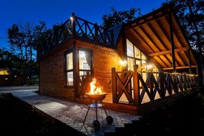 Una cabaña de madera con un ventanal grande, un hoyo para el fuego y un porche cubierto.