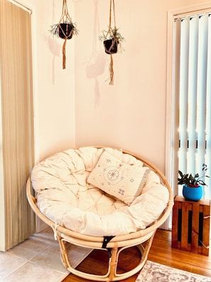 A room with a round, cushioned chair with a decorative pillow, a small table with a blue pot, and two hanging plants.