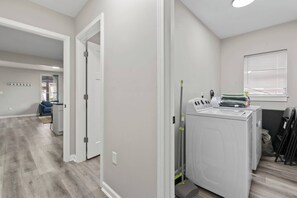A laundry room with a washing machine, dryer, and ironing station. There is a door leading to another room, and a window with blinds.