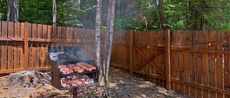 Uma área de churrasco ao ar livre com churrasqueira, lixeira e cerca de madeira.