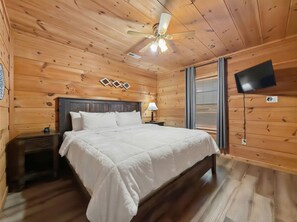 A wooden-paneled room with a bed, a nightstand, a lamp, a ceiling fan, a TV, and a window with blinds. - Sleeps 4, Romantic Chalet, Private Hot Tub, Jacuzzi Tub, Gas Fireplace (Gatlinburg)