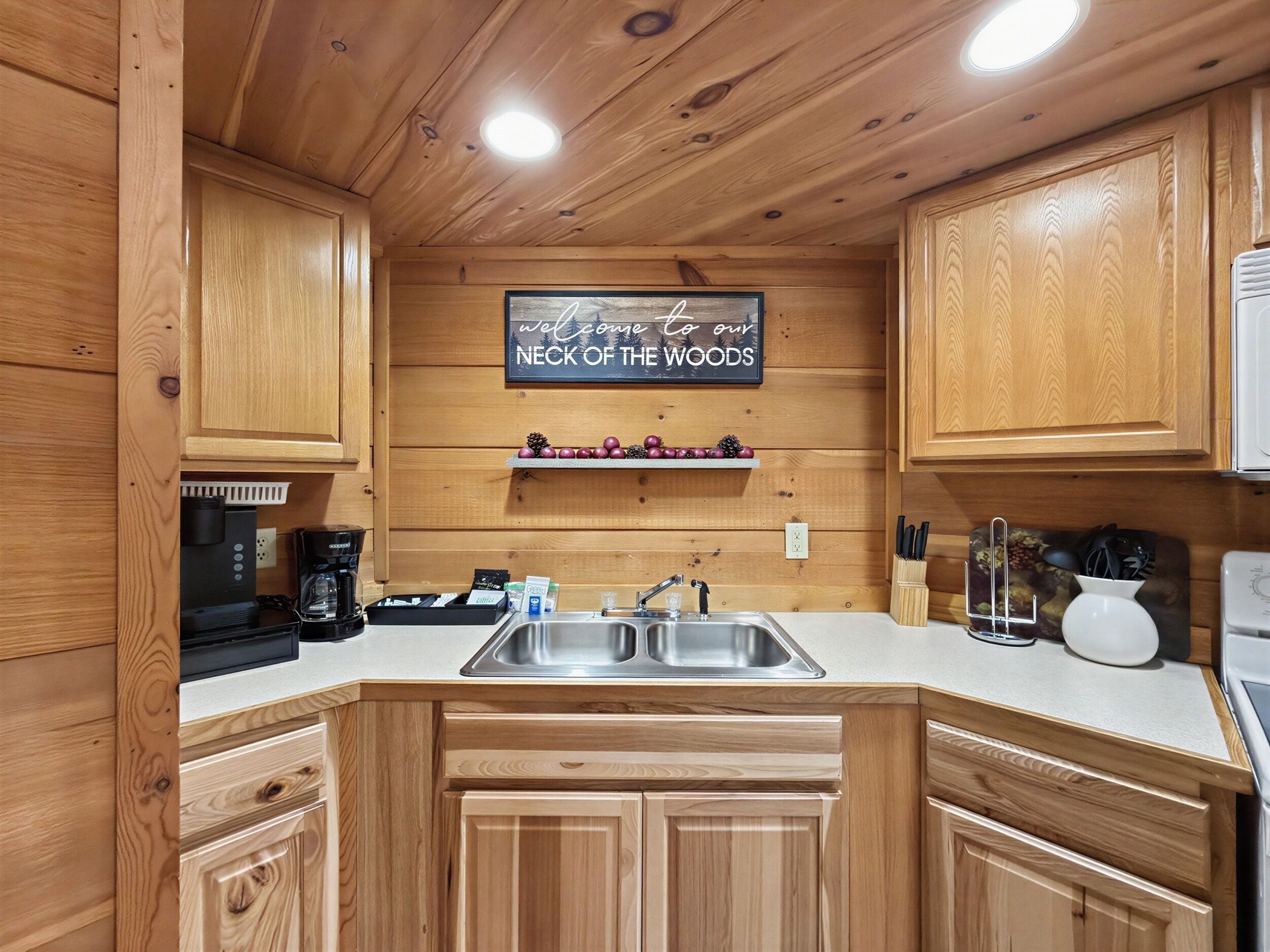 A kitchen with wooden cabinets, a stainless steel sink, and a sign that reads 'Welcome to our NECK OF THE WOODS.'
