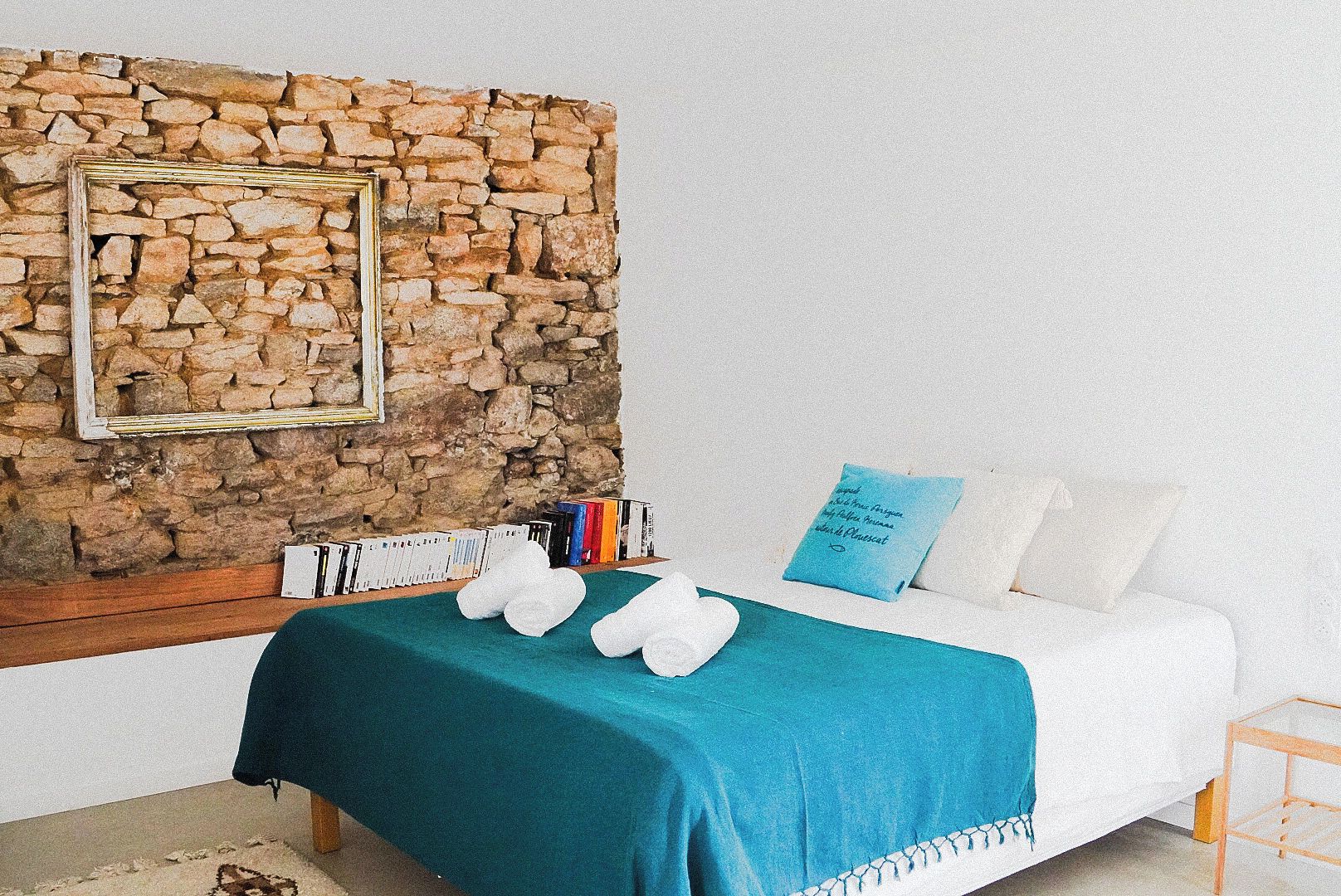 A bedroom with a stone wall, a bed with white bedding, a blue pillow, and a bookshelf with books.