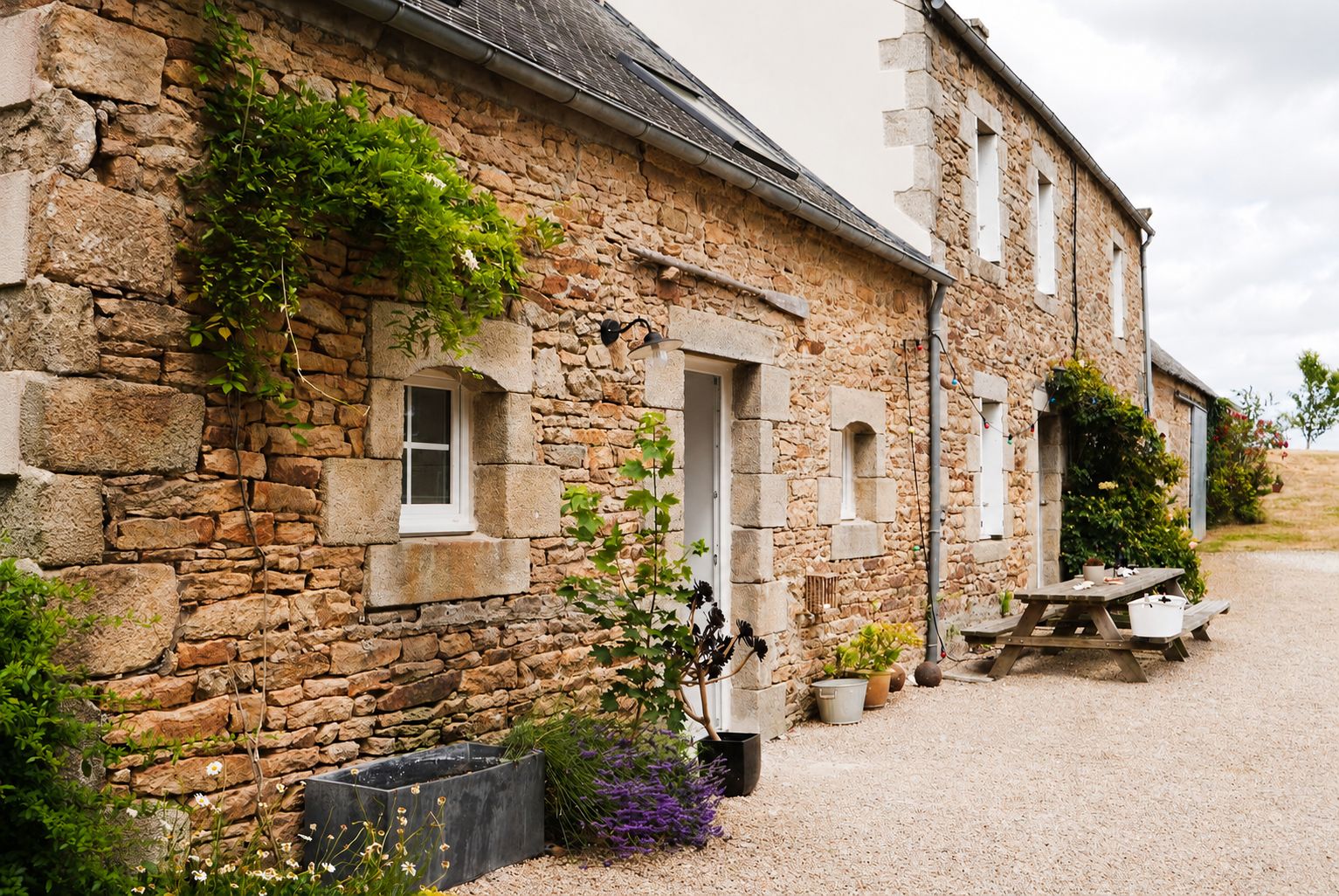 A stone building with a gravel driveway, a wooden bench, and potted plants.