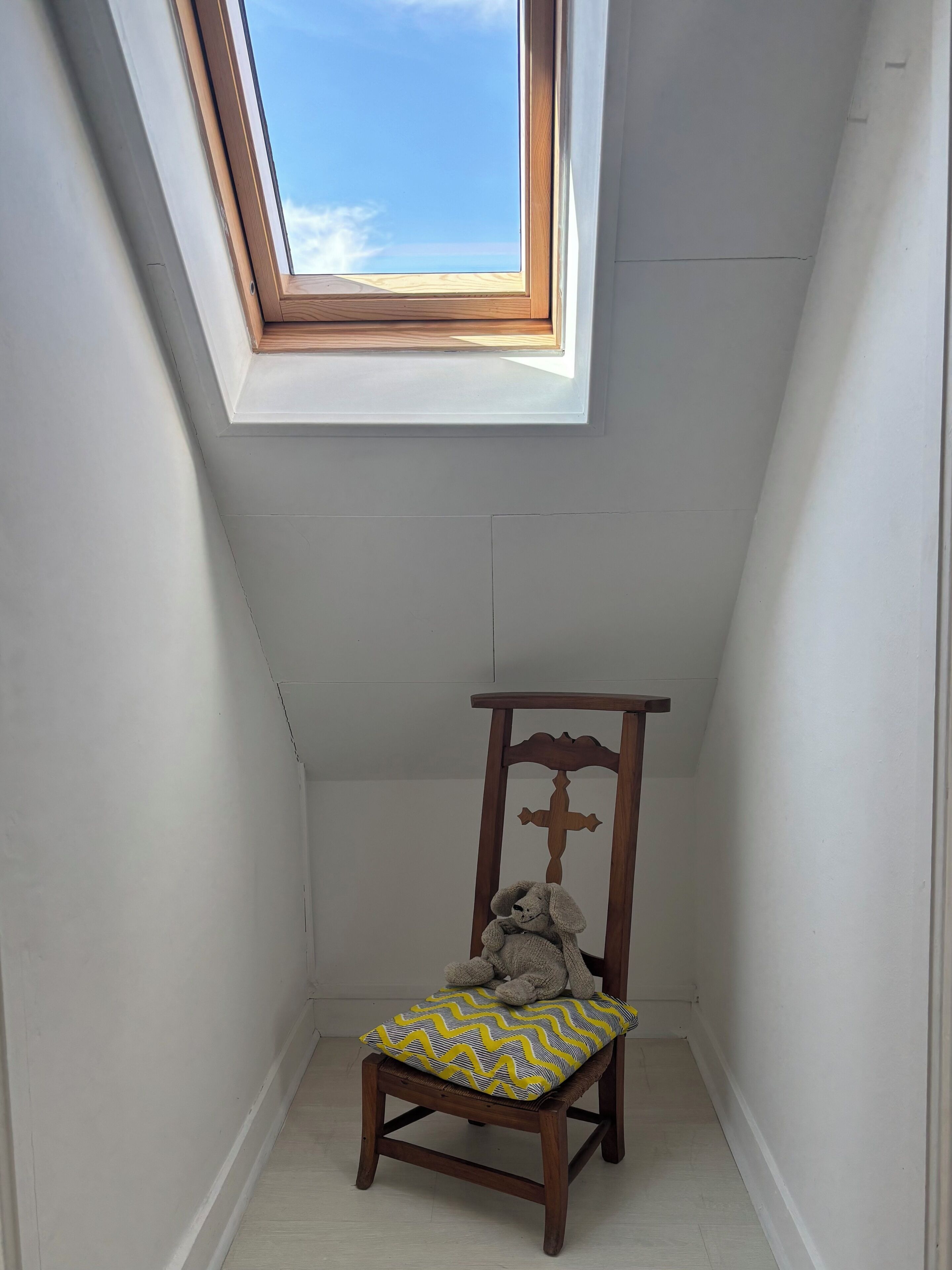 A small room with a sloped ceiling, a wooden chair with a cushion, and a skylight.