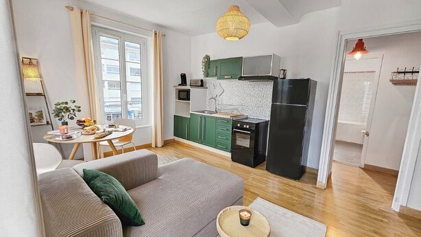 Ett modernt vardagsrum med en soffa, ett litet runt bord med ett ljus och en köksdel med gröna skåp och ett svart kylskåp.