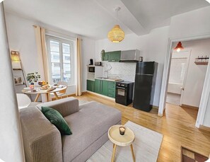 Ein modernes Wohnzimmer mit einer Couch, einem kleinen runden Tisch mit Kerze sowie einer Küchenzeile mit grünen Schränken und einem schwarzen Kühlschrank.