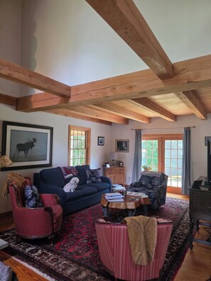 Una sala de estar con techo alto, vigas de madera, un sofá, un sillón rojo, un tapete estampado y una mesita baja con libros y una planta.