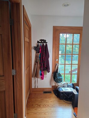 Ein Korridor mit Holzboden, einem Kleiderständer mit verschiedenen Kleidungsstücken, einem Fenster mit Blick ins Grüne und einer Tür mit hölzernem Rahmen.