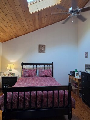 Un dormitorio con cama, mesita de noche, lámpara, ventilador de techo y un cuadro en la pared.