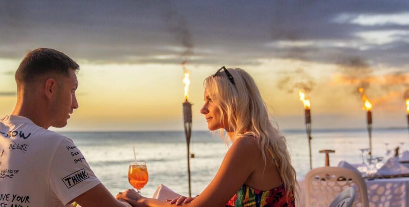 Una pareja cenando en una mesa con vista al océano y al atardecer.