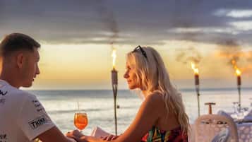Una pareja cenando en una mesa con vista al océano y al atardecer.