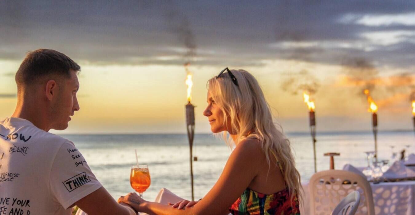 A couple seated at a table with a view of the sea and a sunset in the background.