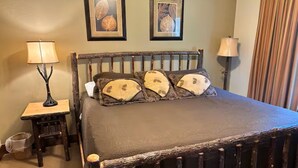 Ein Bett mit dunklem Holzgestell und grauem Lattenrost, flankiert von zwei Nachttischen mit Lampen, in einem Zimmer mit beige gestrichener Wand und Wandbild mit Blättern.