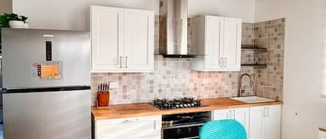 A modern kitchen with white cabinets, a stainless steel refrigerator, a gas stove, and a wooden dining table with blue chairs.