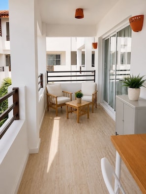 Uma varanda com mesa e cadeiras de madeira, um vaso de planta e vista do interior do edifício.