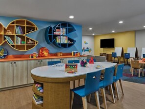 Zona de juegos infantiles con una mesa redonda, sillas azules y dos grandes estanterías llenas de juguetes y libros.