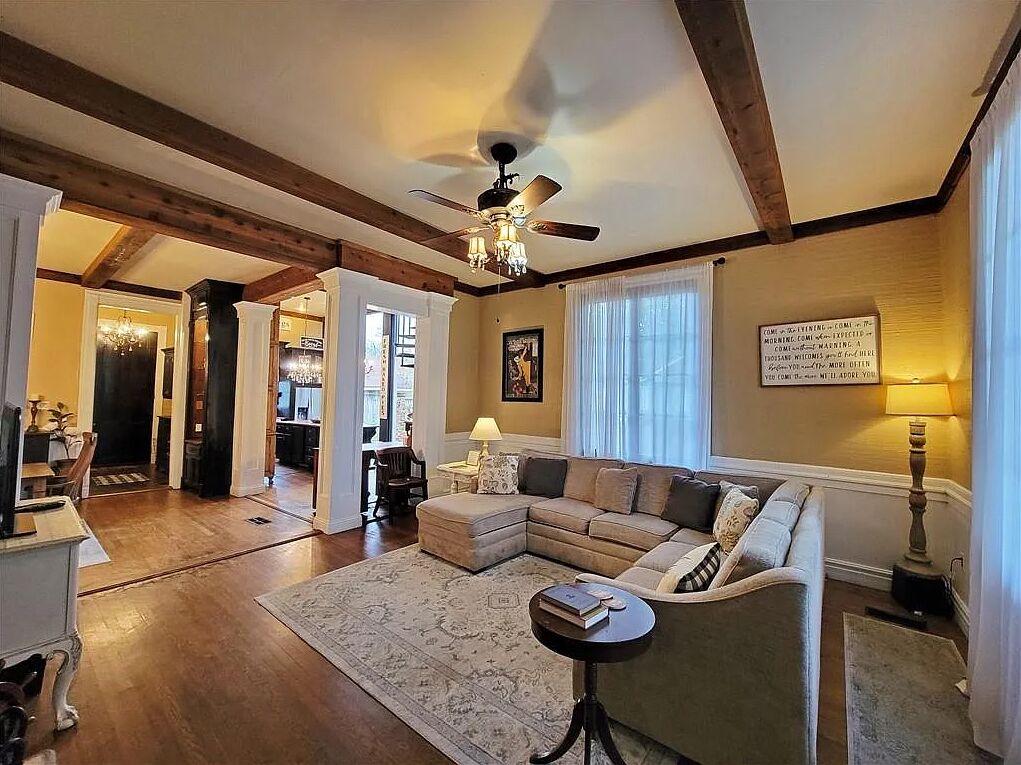 A spacious living room with a large sectional sofa, a round coffee table, and a ceiling fan. There is a dining area visible in the background.