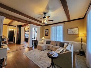A spacious living room with a large sectional sofa, a round coffee table, and a ceiling fan. There is a dining area visible in the background. - The Steamboat House, Captain's Quarters-Bed & Breakfast Style Experience (Natchitoches)