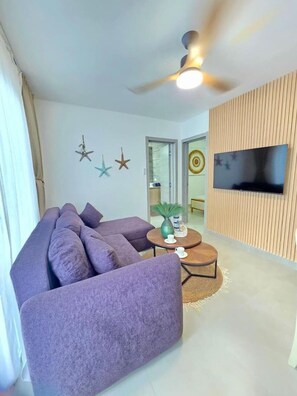A living room with a purple sofa, a round coffee table, a ceiling fan, and a flat-screen TV mounted on a wooden panel wall. - ANYA 5 Chic Coastal Oasis • King Bed & Balcony (Bayahíbe)
