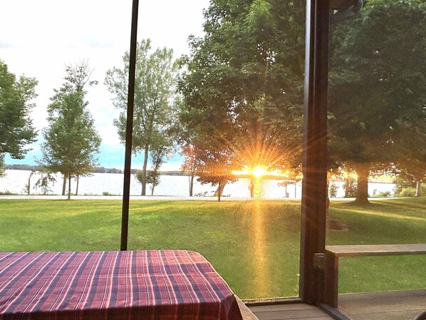 A room with a view of a grassy area and trees, a table with a striped tablecloth, and a window with a view of the sunset over a body of water.
