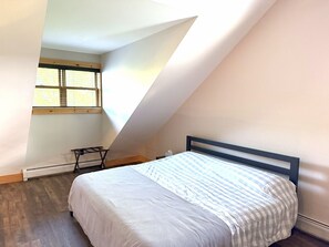 Ein Schlafzimmer mit schrägem Dach, einem Bett mit gestreifter Bettdecke, einem Fenster mit Jalousien und einer Heizung.