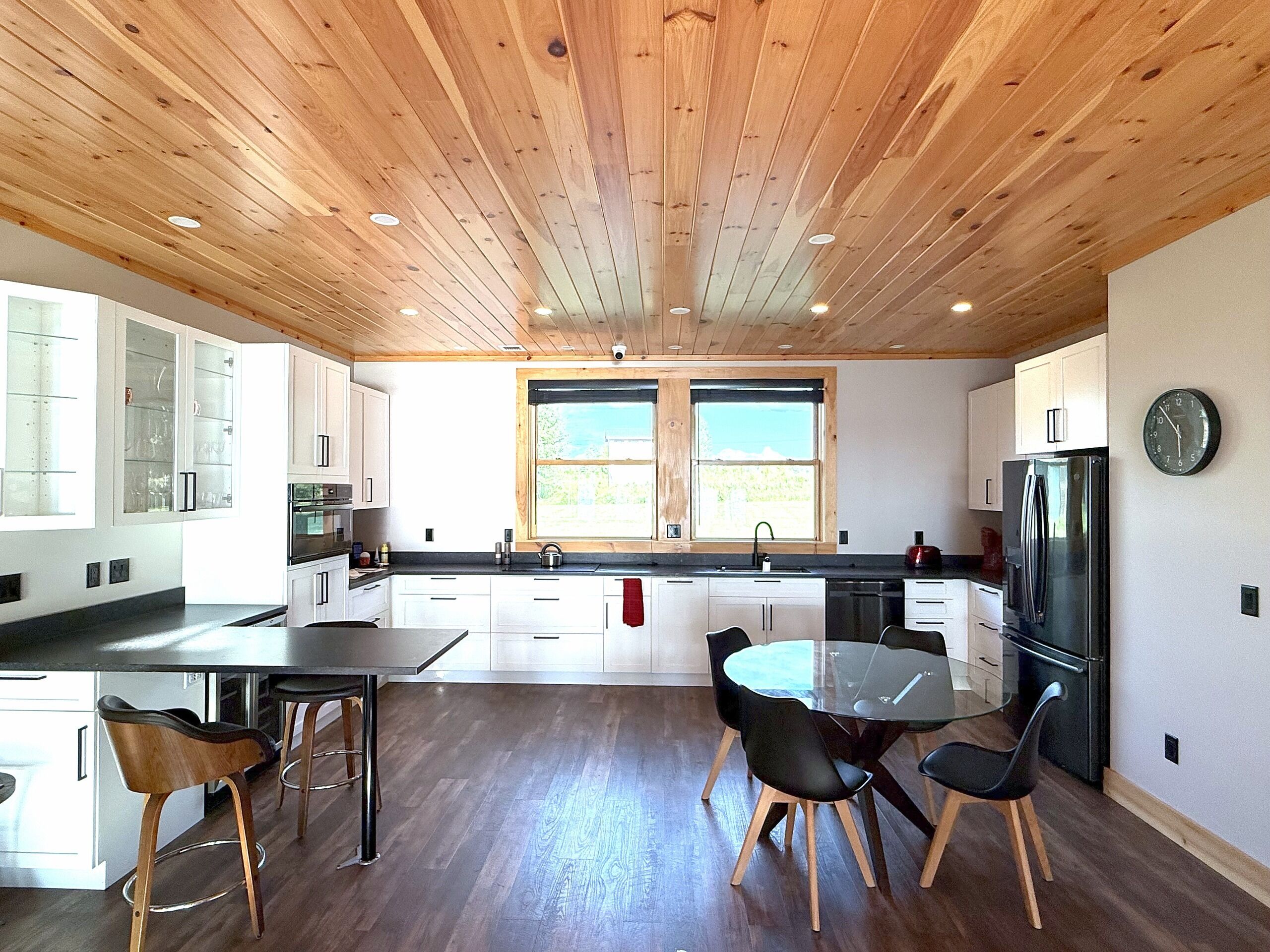 A modern kitchen with wooden ceiling, white cabinets, a glass dining table, and black chairs.